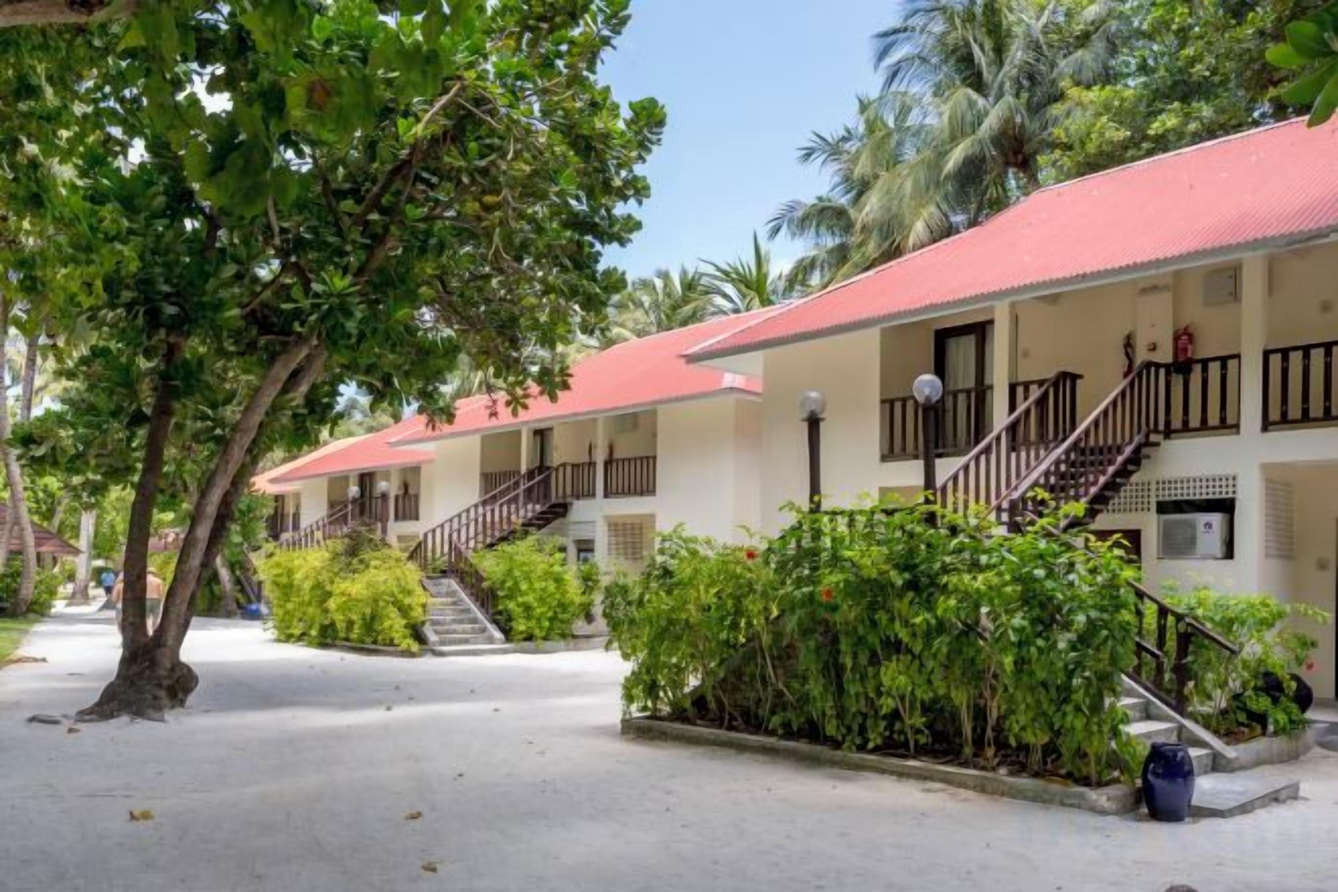 A row of two-story buildings with balconies, surrounded by greenery and palm trees.