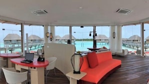 Un área de descanso moderna con un sofá rojo, una laptop sobre una mesita y vistas a bungalows sobre el agua y un mar azul cristalino.
