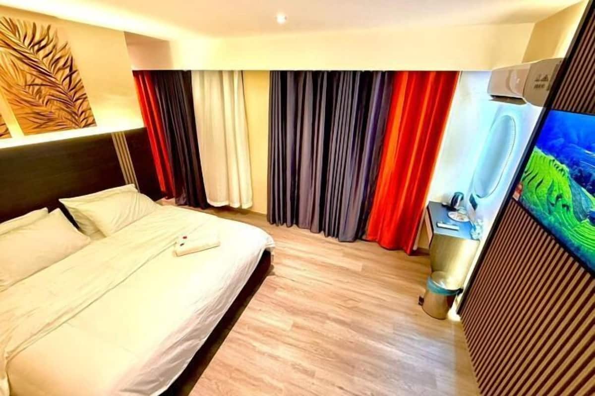 A hotel room with a bed, curtains, a TV, and a desk with a trash bin.