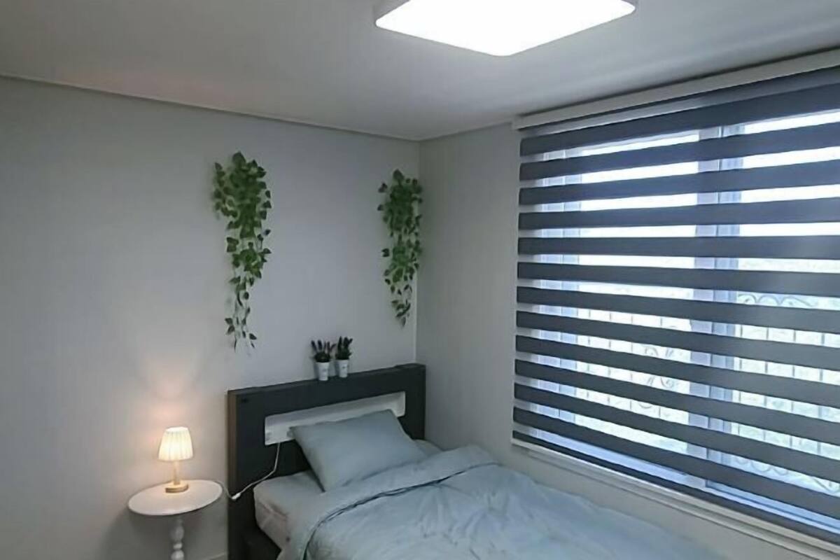 Un dormitorio con cama, mesita de noche con lámpara, ventana con persianas y dos plantas colgantes.