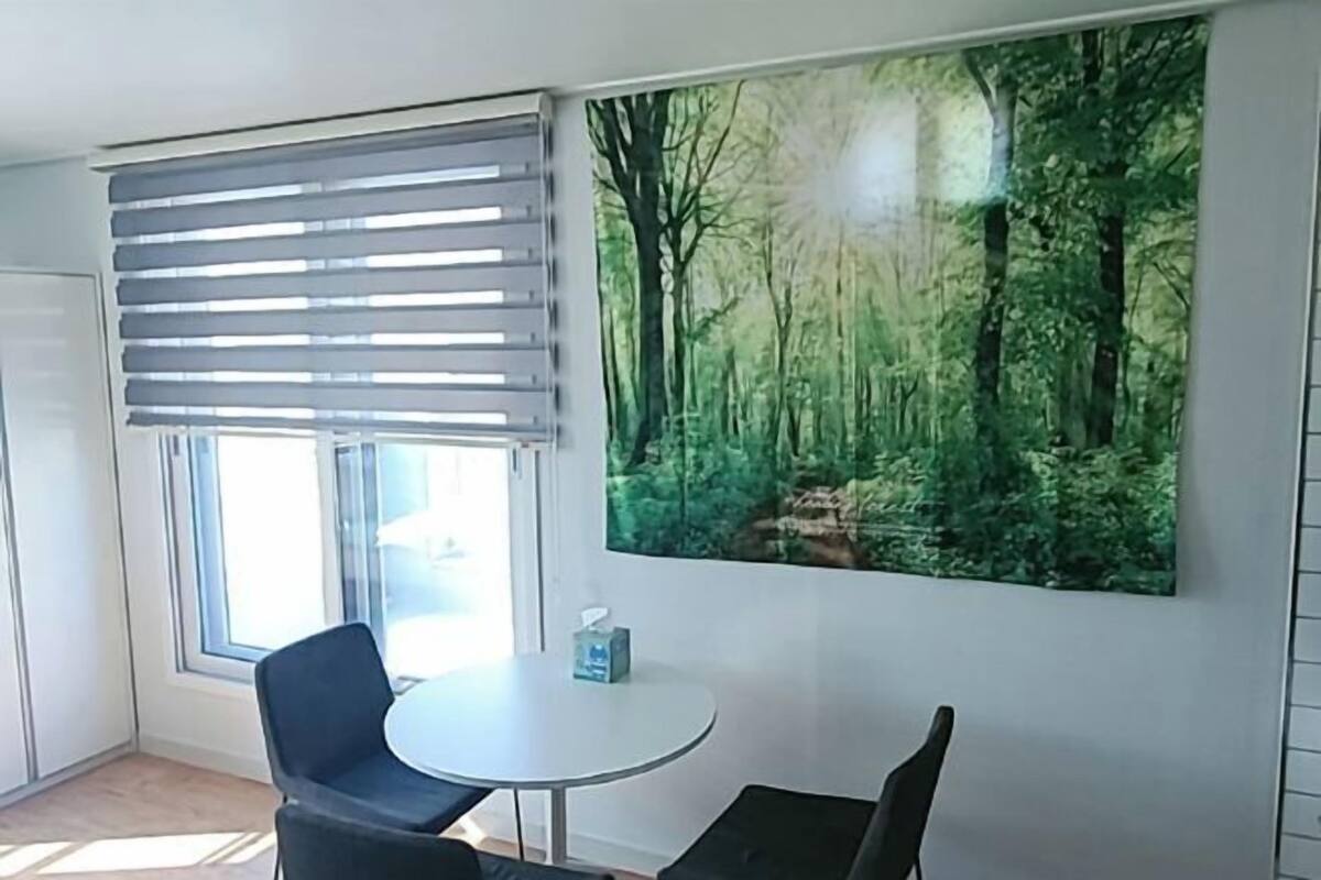 Una habitación con una mesa redonda, dos sillas, un ventanal con persianas y un mural grande con motivos forestales.