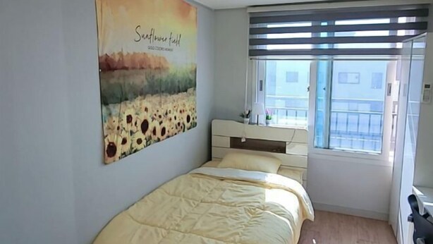 A bedroom with a bed, a window with blinds, a wall with a floral painting, and a small bedside table. - Incheon Airport ANN Guesthouse (Incheon)