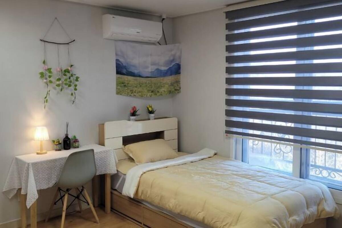 Un dormitorio con cama, mesita de noche, lámpara, equipo de aire acondicionado, ventana con persianas y una planta en la pared.
