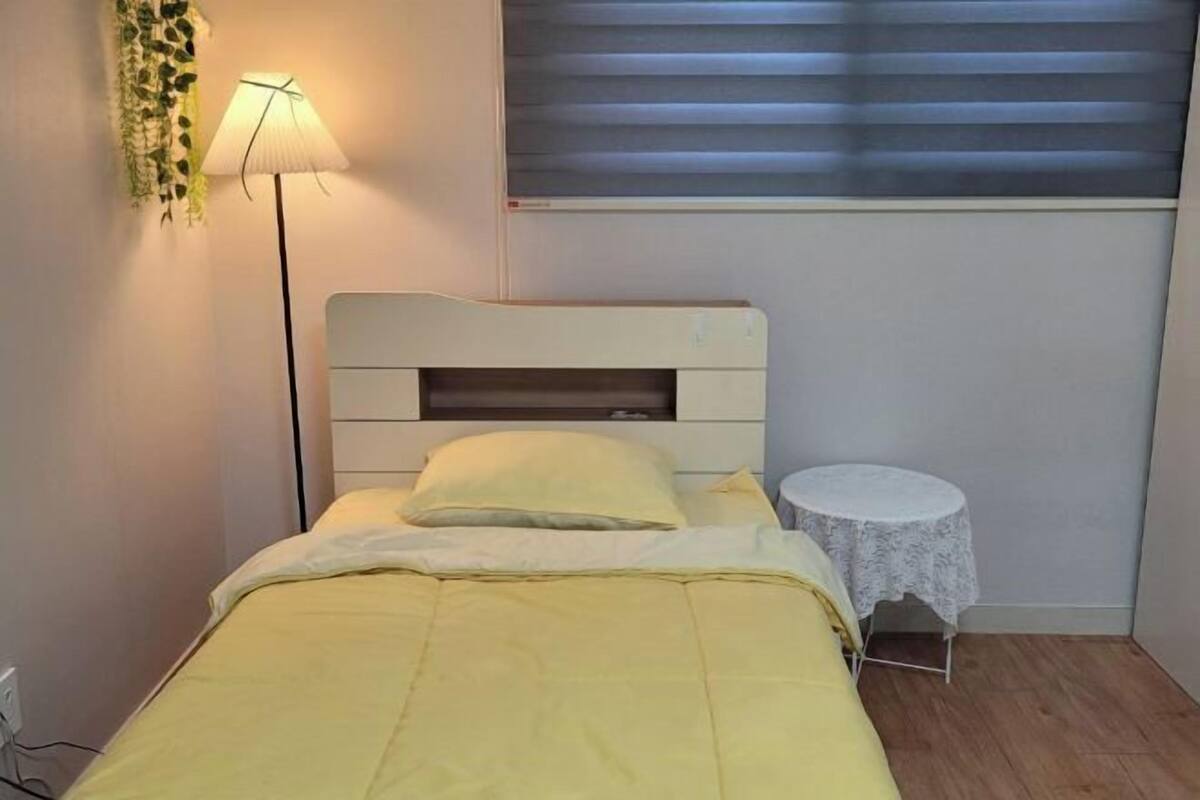 Una cama con cabecera blanca, una mesita de noche con mantel de encaje y una planta colgante.