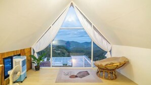 A room with a large window, a fan, a chair, a small table, and a potted plant. - Phutawan monjam (Mae Rim)