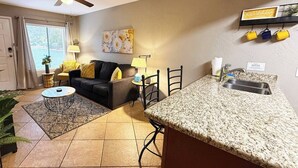 Een woonkamer met een bank, salontafel en stoelen, een keuken met granieten aanrecht en een spoelbak, en een plafondventilator.