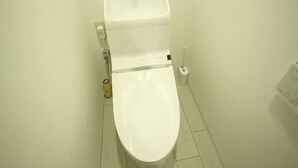 Μια λευκή τουαλέτα σε ένα μπάνιο με έναν απλό τοίχο και έναν μικρό κάδο απορριμμάτων δίπλα της.
