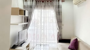 A living room with a sofa, a TV on a stand, a window with sheer curtains, and a wall-mounted air conditioner. - Daily Apartment & Residence (Phnom Penh)