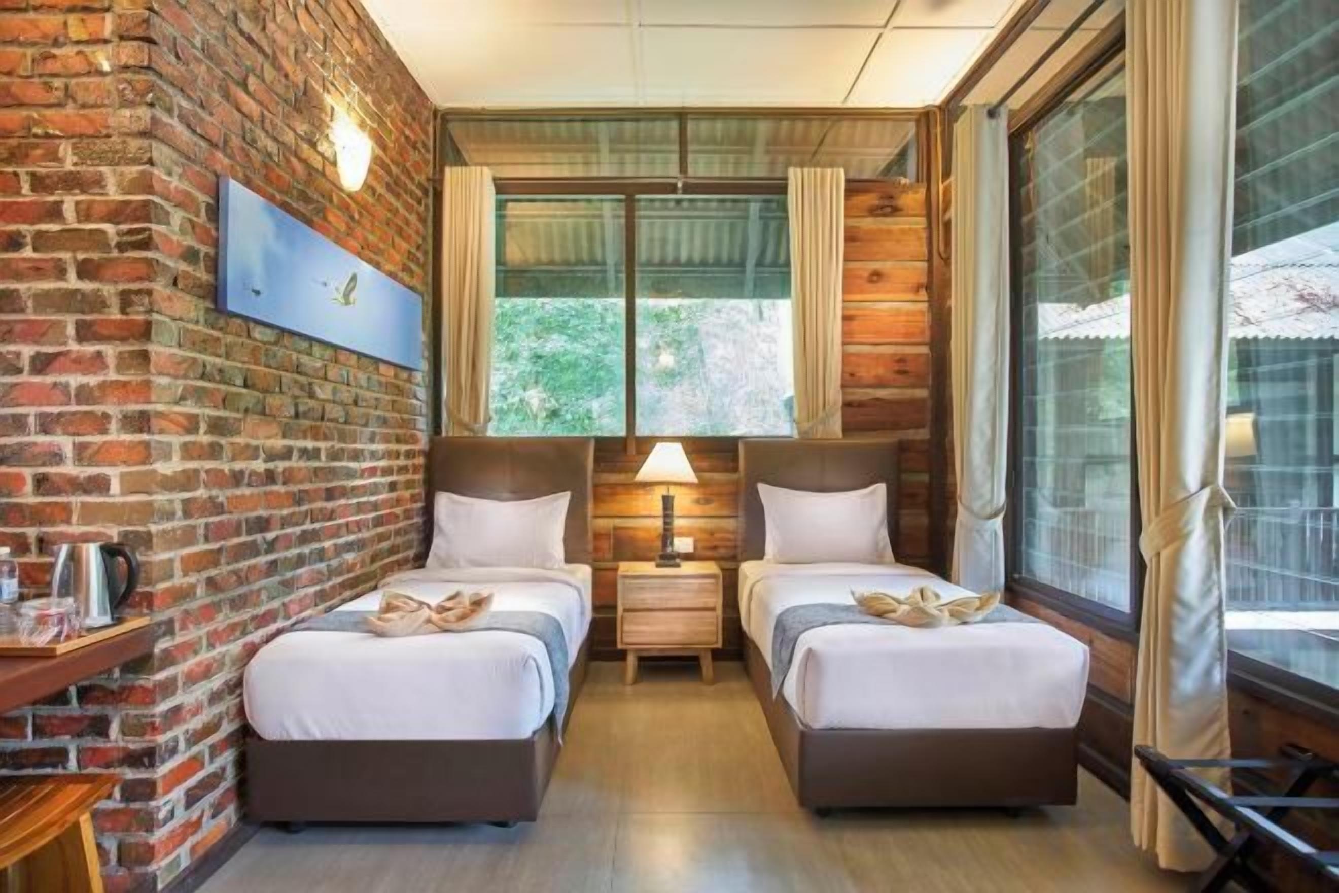 A hotel room with two beds, a brick wall, a window with curtains, and a nightstand with a lamp.