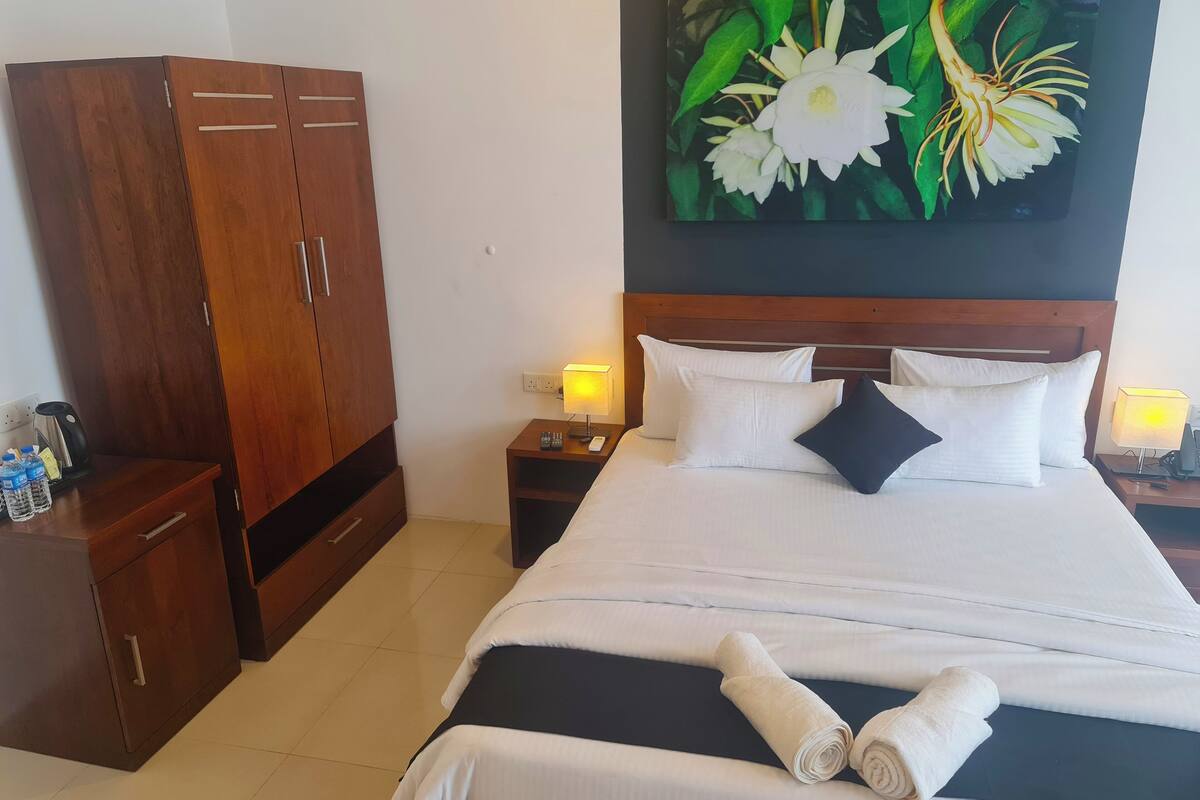 Une chambre d’hôtel avec une armoire en bois, un lit avec des oreillers blancs et foncés, une table de chevet avec une lampe et un tableau représentant des fleurs au mur.