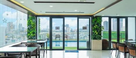 A modern hotel room with a large window, a dining area with tables and chairs, and a view of the cityscape.