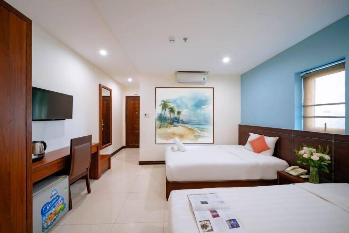 Habitación de hotel con dos camas, un escritorio con televisor, un cuadro en la pared y una ventana con vistas.