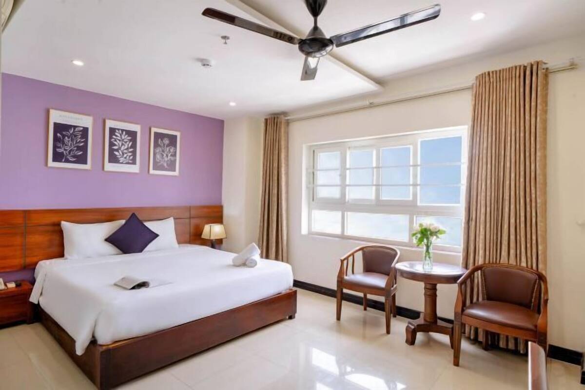 Un cuarto de hotel con una cama grande, un ventilador de techo, una ventana con cortinas y obras de arte enmarcadas en la pared.