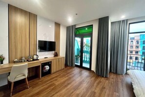 A modern hotel room with a wooden desk, a flat-screen TV, a bed, and a balcony with a view of buildings. - Homestay SWAN BY KAT (Nghia Tru)