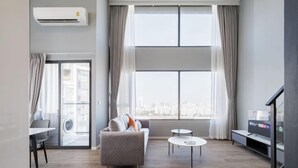 A modern living room with a sofa, coffee table, and TV stand, large windows with curtains, and an air conditioning unit. - Loft Apartments in Bangkok near Jodd Night Market [Nearly 711+ Central Pharam 9 Shopping Center] (Bangkok)