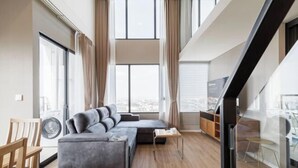 A modern living room with a grey sectional sofa, a small table, and a view of the city through large windows. - Loft Apartments in Bangkok near Jodd Night Market [Nearly 711+ Central Pharam 9 Shopping Center] (Bangkok)