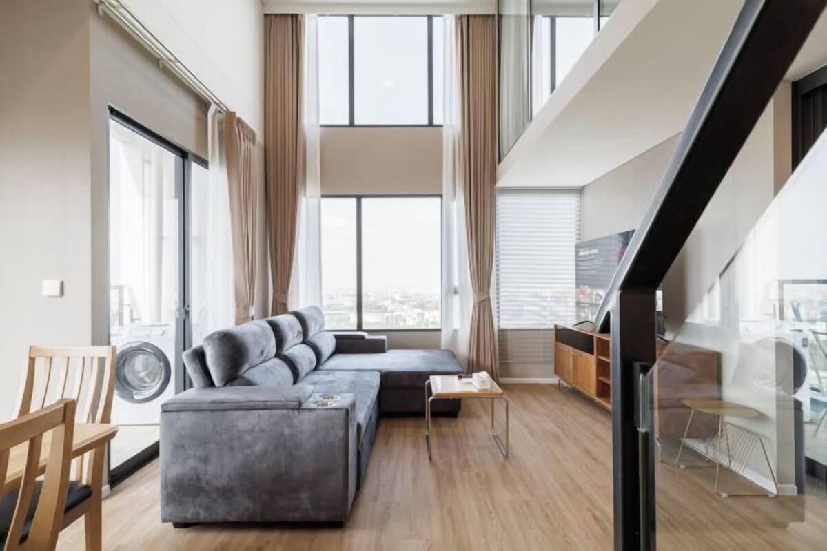 Ein modernes Wohnzimmer mit einem grauen Ecksofa, einem kleinen Tisch und Blick auf die Stadt durch große Fenster.