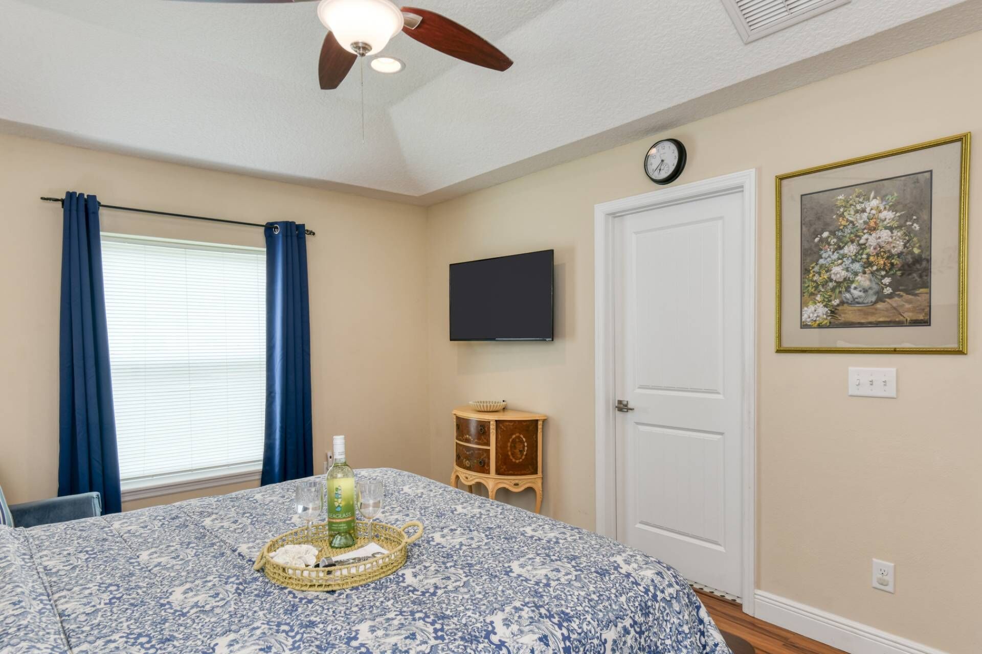 Une chambre à coucher avec un lit, un ventilateur de plafond, une fenêtre avec des rideaux, une table de chevet avec une bouteille et des verres, une horloge et un tableau encadré au mur.