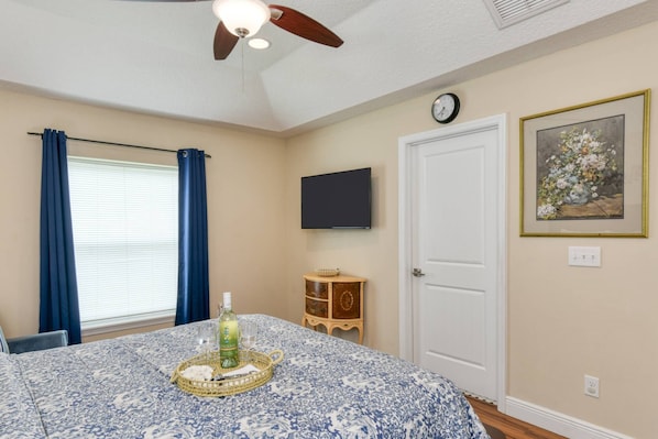 A bedroom with a bed, a ceiling fan, a window with curtains, a bedside table with a bottle and glasses, a clock, and a framed picture on the wall.