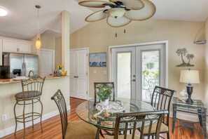 A dining area with a glass table surrounded by chairs, a ceiling fan, and a kitchen in the background.