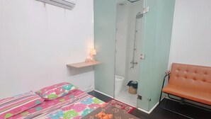 Een kleine kamer met een bed, een bankje, een klein tafeltje met een lamp en een douchegedeelte.