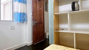 Een kamer met een houten deur, een raam met blauwe gordijnen, een witte plank met een waterkoker en een witte stoel.