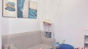 Een kamer met een beige bank, een blauwe stoel en een kleine tafel met een plant. Er hangen drie afbeeldingen in lijsten aan de muur.