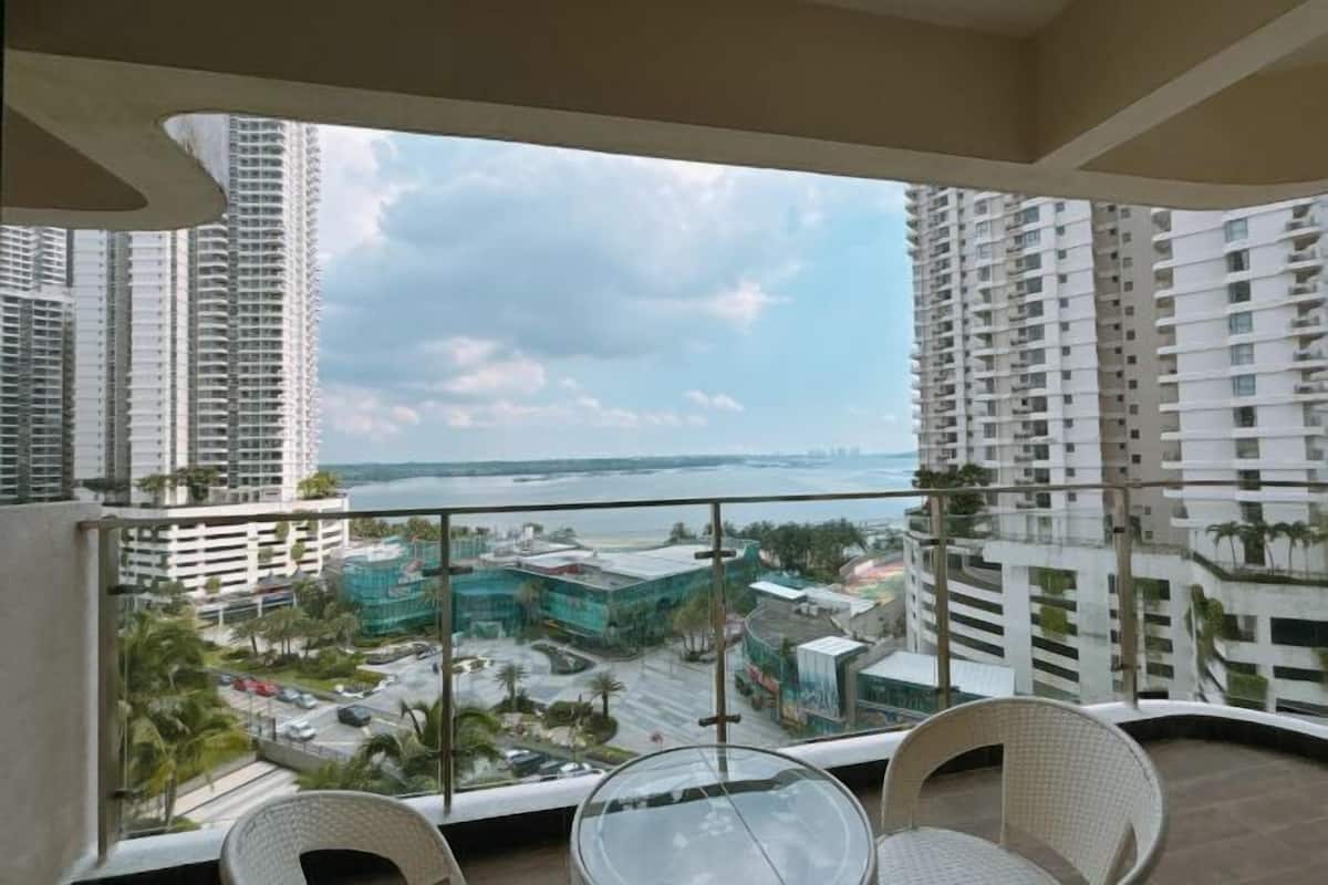 A balcony with a glass table and two wicker chairs, overlooking a cityscape with high-rise buildings and a body of water.