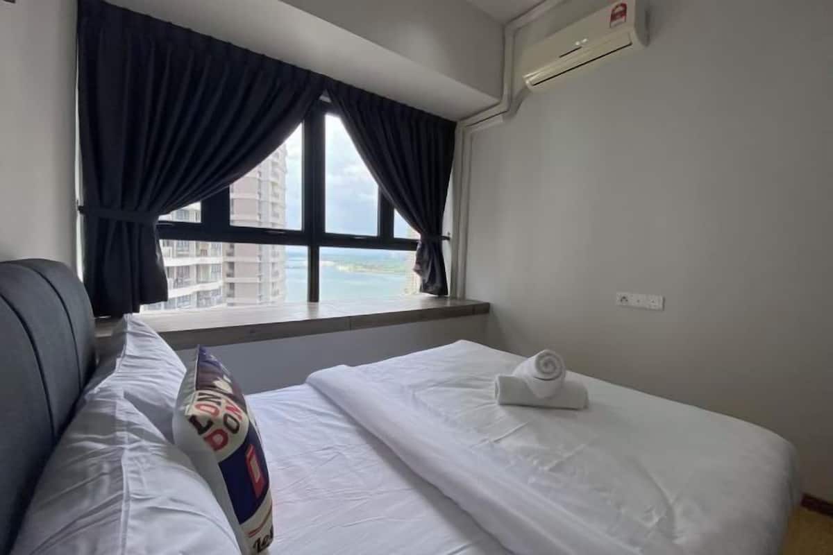 A hotel room with a bed, a window with curtains, an air conditioning unit, and a view of buildings.