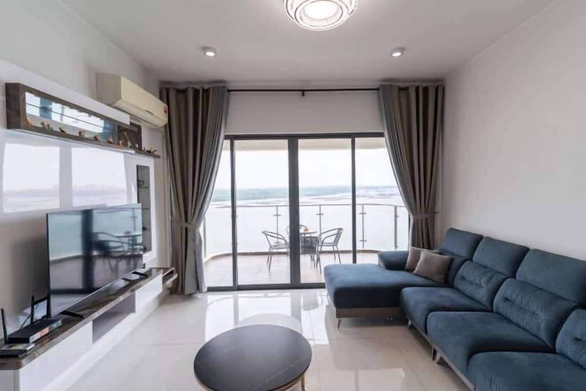 A modern living room with a large flat-screen TV, a blue sofa, a round coffee table, and a balcony with a view.