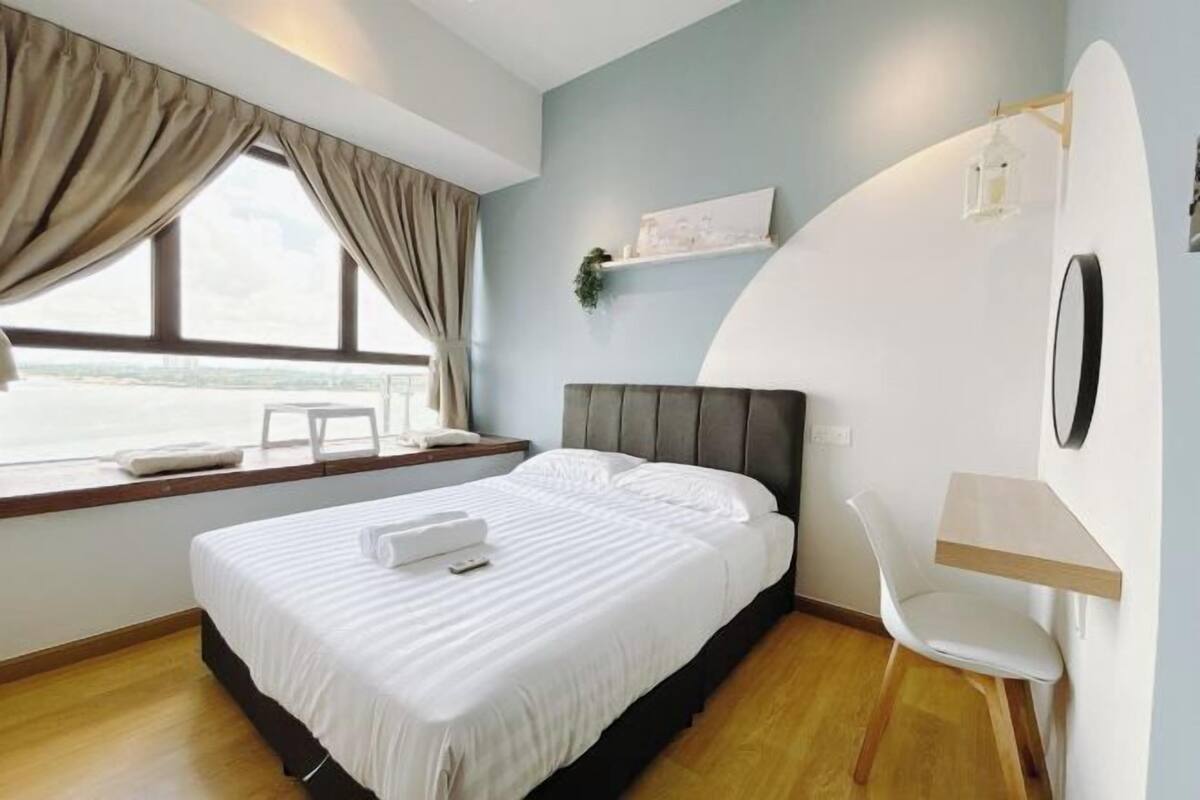 A hotel room with a bed, a desk, a chair, a window with curtains, and a shelf with a plant.