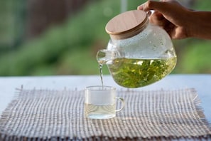 A hand pouring tea from a glass pitcher into a cup on a woven mat. - Bon r kaad (Mae Rim)