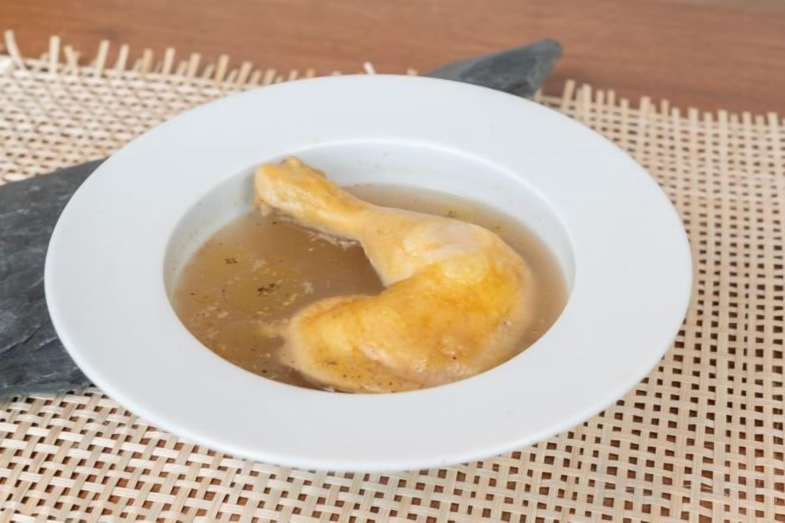 Un tazón blanco con una pieza de pollo frito en un líquido, posiblemente caldo, sobre un tapete texturizado.