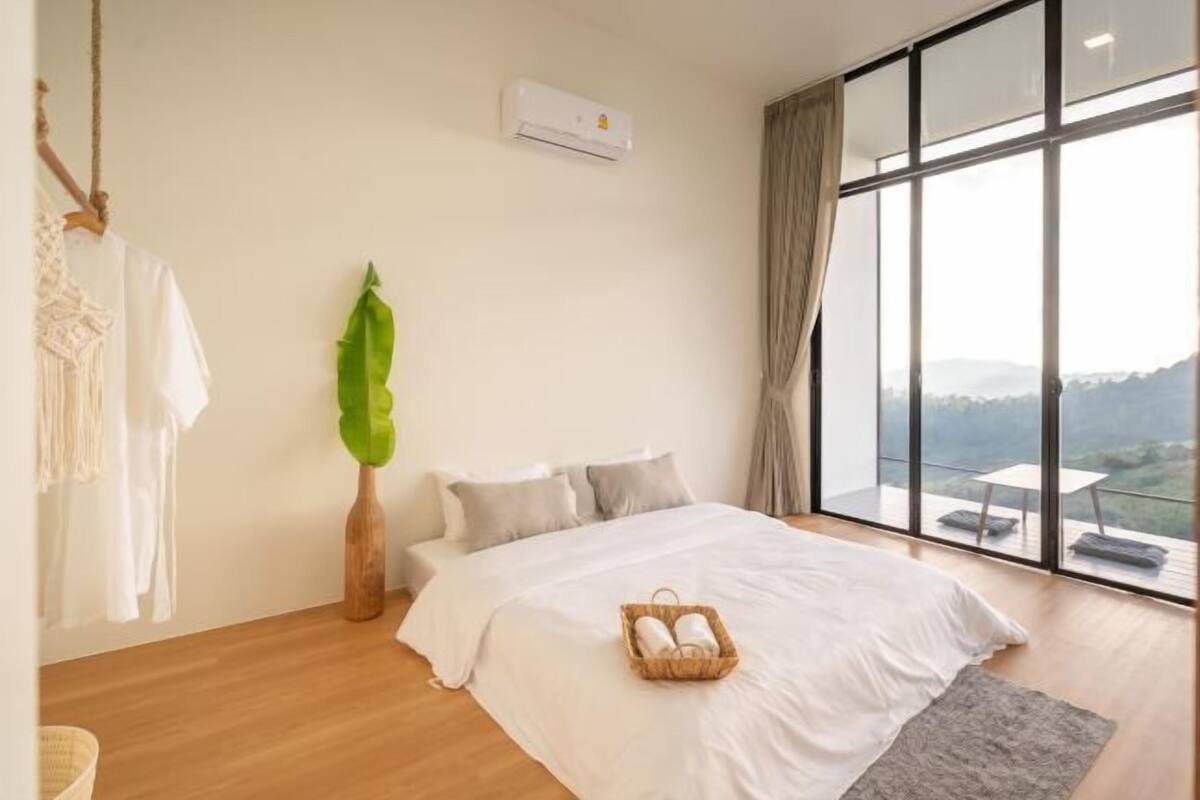 A bedroom with a large bed, a view of the outdoors, and a hanging plant.