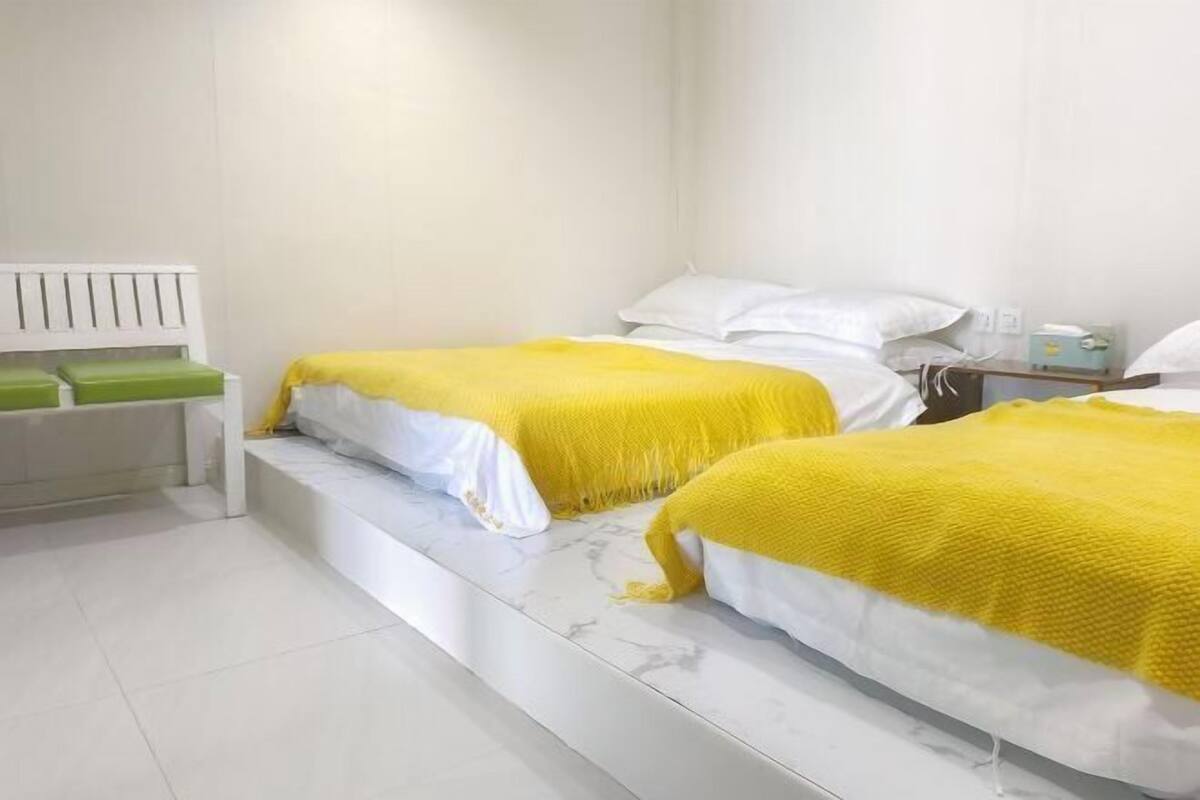 Une chambre avec deux lits, chacun doté d’un matelas blanc et d’une couverture jaune. On y trouve un banc blanc avec des coussins verts, une tête de lit blanche et une petite table de chevet avec une lampe.