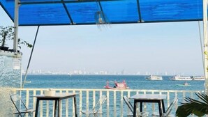 A covered outdoor seating area with tables and chairs, overlooking a body of water with boats and a distant city skyline. - Rock Resort Koh Larn (Koh Larn)