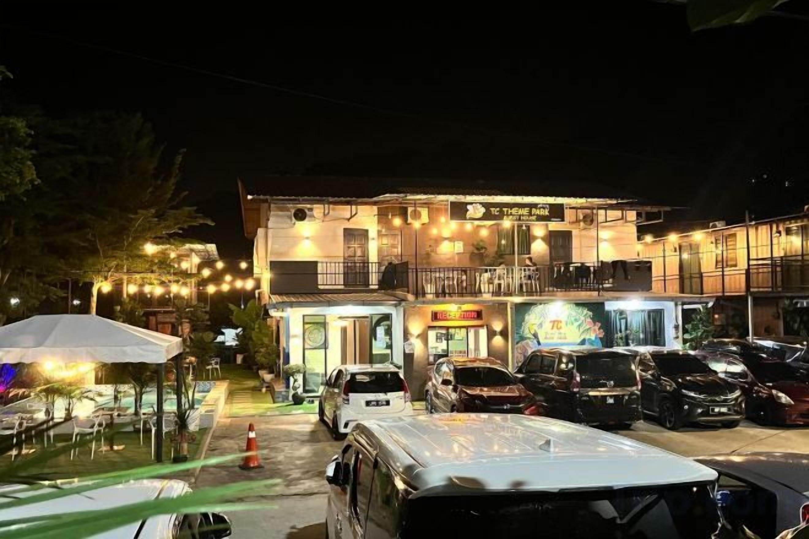 A two-story building with a balcony, a parking area with several cars, and outdoor seating with umbrellas.