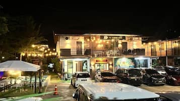 A two-story building with a balcony, a parking area with several cars, and outdoor seating with umbrellas.