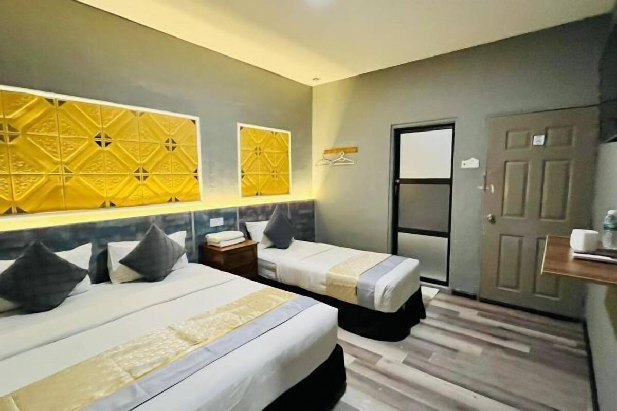 Habitación de hotel con dos camas, un cabecero de madera, una mesita de noche pequeña y un estante en la pared, además de una puerta.