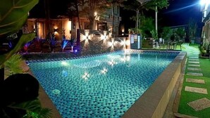 Ein Schwimmbecken mit Kachelmuster, umgeben von einem gut beleuchteten Gehweg und Grünflächen.