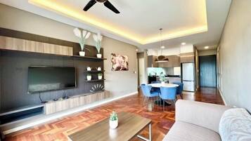 A modern living room with a flat-screen TV, wooden flooring, a ceiling fan, and a dining area with blue chairs.