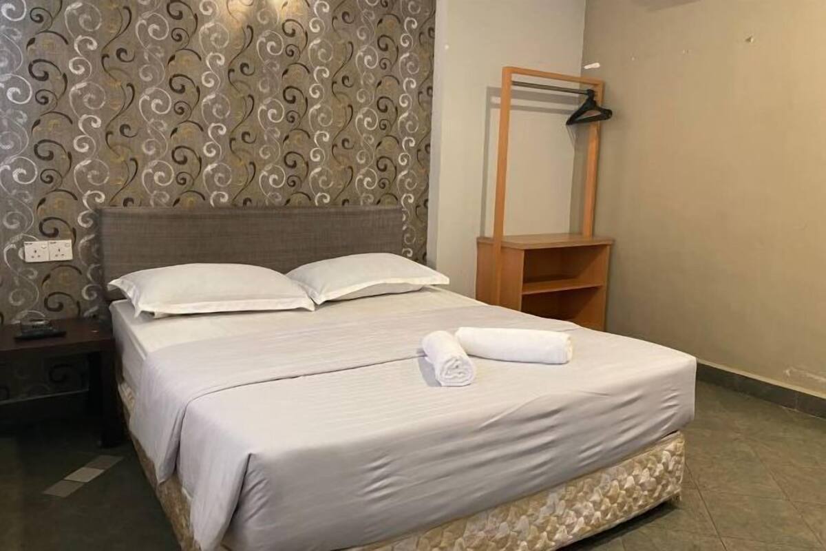 Un lit bien fait, avec du linge de lit blanc et une serviette pliée, une table de chevet en bois, un miroir au mur et un papier peint à motifs.