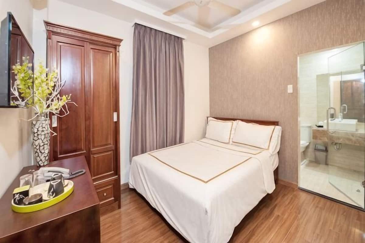 A hotel room with a bed, a desk with a vase, a wardrobe, and a bathroom with a glass shower.