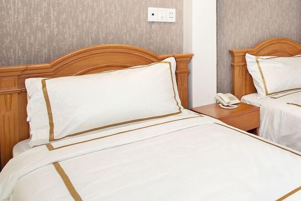 Une chambre d’hôtel avec deux lits, chacun doté de draps blancs et d’une bordure dorée, d’une tête de lit en bois et d’un téléphone sur la table de chevet.