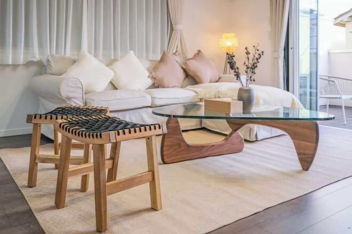 A modern living room with a glass coffee table, two wooden stools, a white sofa with cushions, and a vase with branches.