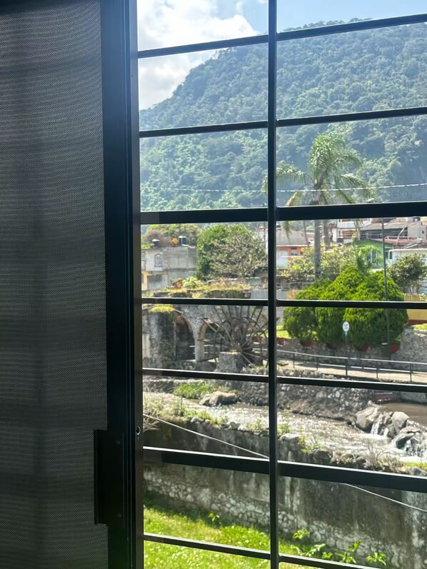 A view through a window with metal bars, featuring a stone water wheel, greenery, and a mountainous backdrop.