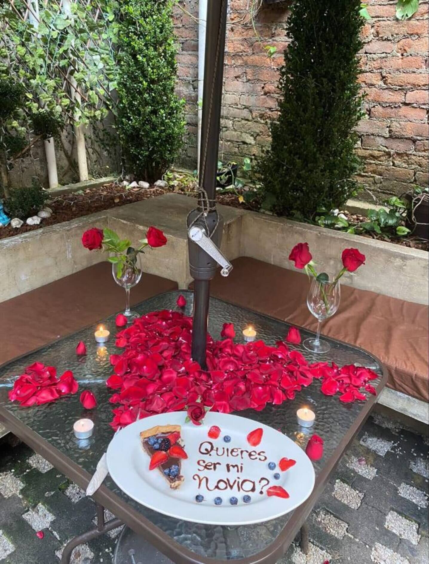 Tavolo da esterno per mangiare con petali di rosa, candele e un piatto di dessert con una scritta.
