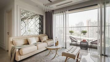 Una sala de estar moderna con sofá, mesa redonda de centro y reposapiés, ventanas amplias con cortinas, y vista a la ciudad.