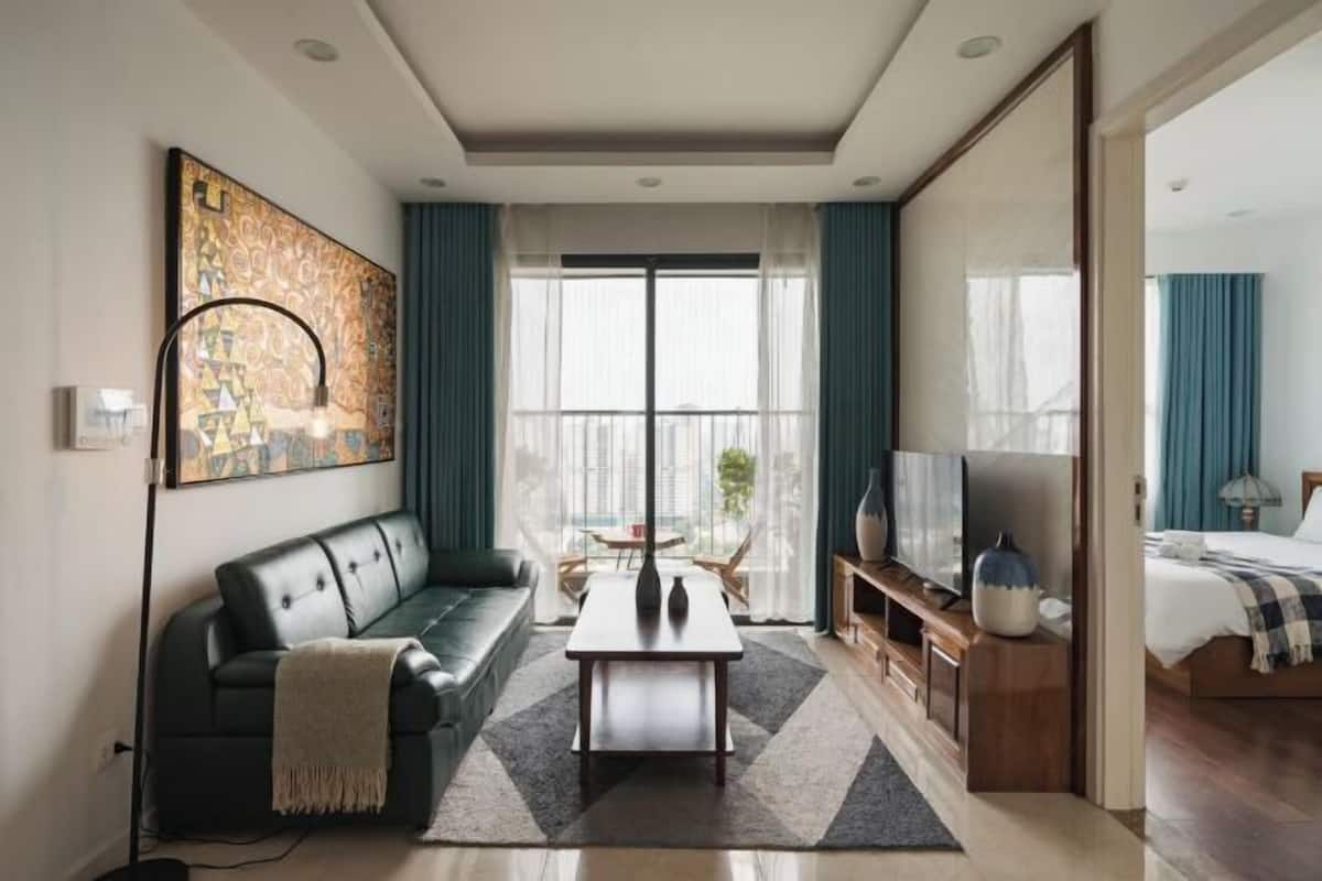 Ein modernes Wohnzimmer mit einer Couch, einem Couchtisch und einem Fernseher. Ein großes Fenster mit Vorhängen, ein Gemälde an der Wand und ein Balkon, der durch das Fenster sichtbar ist.
