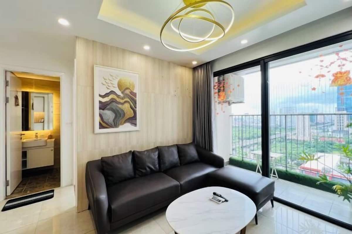 Ein modernes Wohnzimmer mit einer schwarzen Ledersofa, einem runden Couchtisch, einem Gemälde an der Wand und einem großen Fenster mit Blick auf Gebäude und Grünflächen.